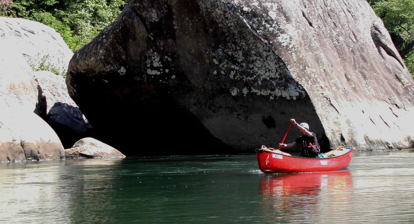 A person paddles a red canoe on still water beside a large boulder 